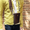A person wearing a yellow shirt and dark pants is standing in front of a brick wall. The person is wearing a brown leather crossbody bag with an adjustable strap.