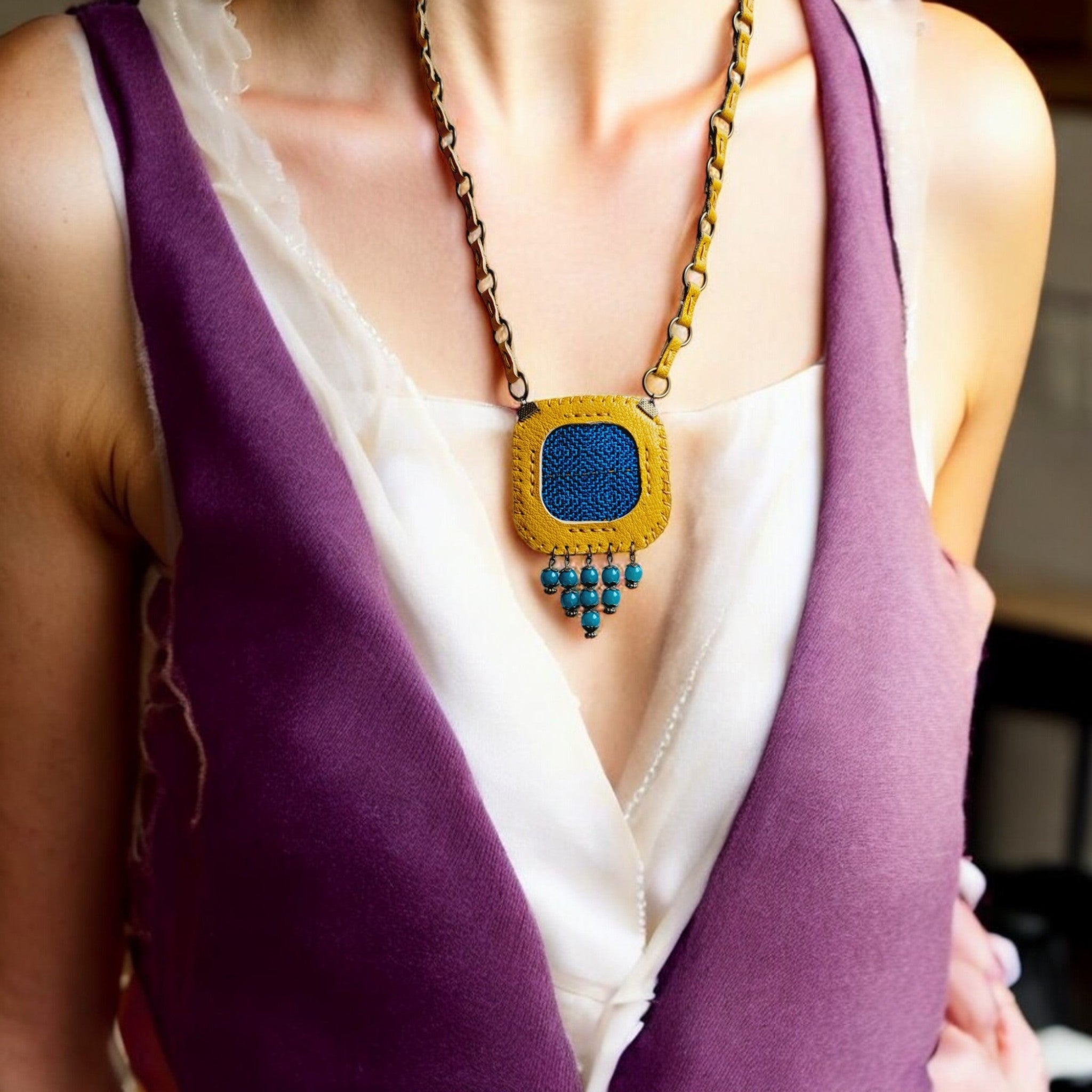 A person wearing a purple and white garment showcasing a handmade necklace with a blue woven square pendant and teal bead accents.