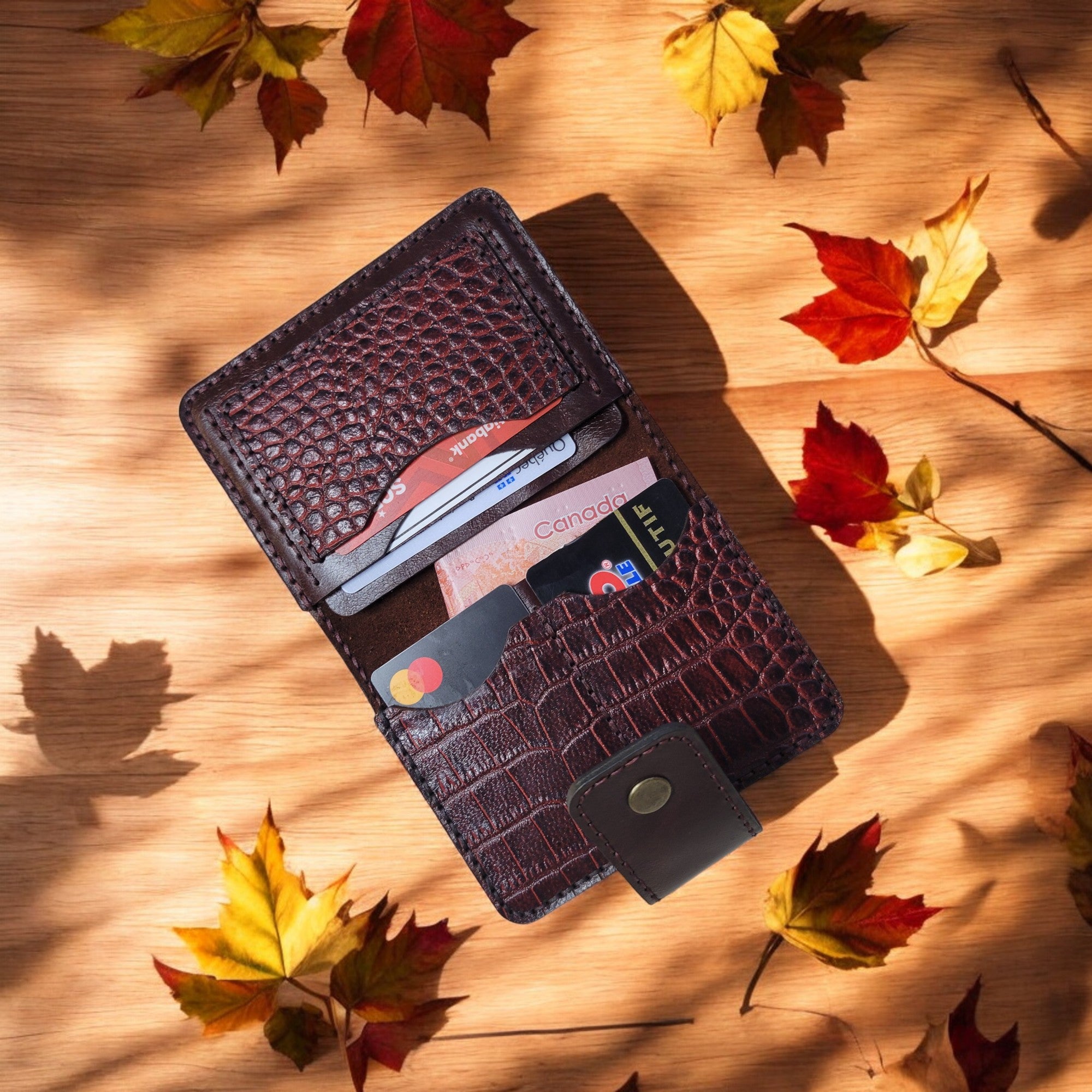 A brown leather wallet with a crocodile pattern displayed on a surface with autumn leaves scattered around it.