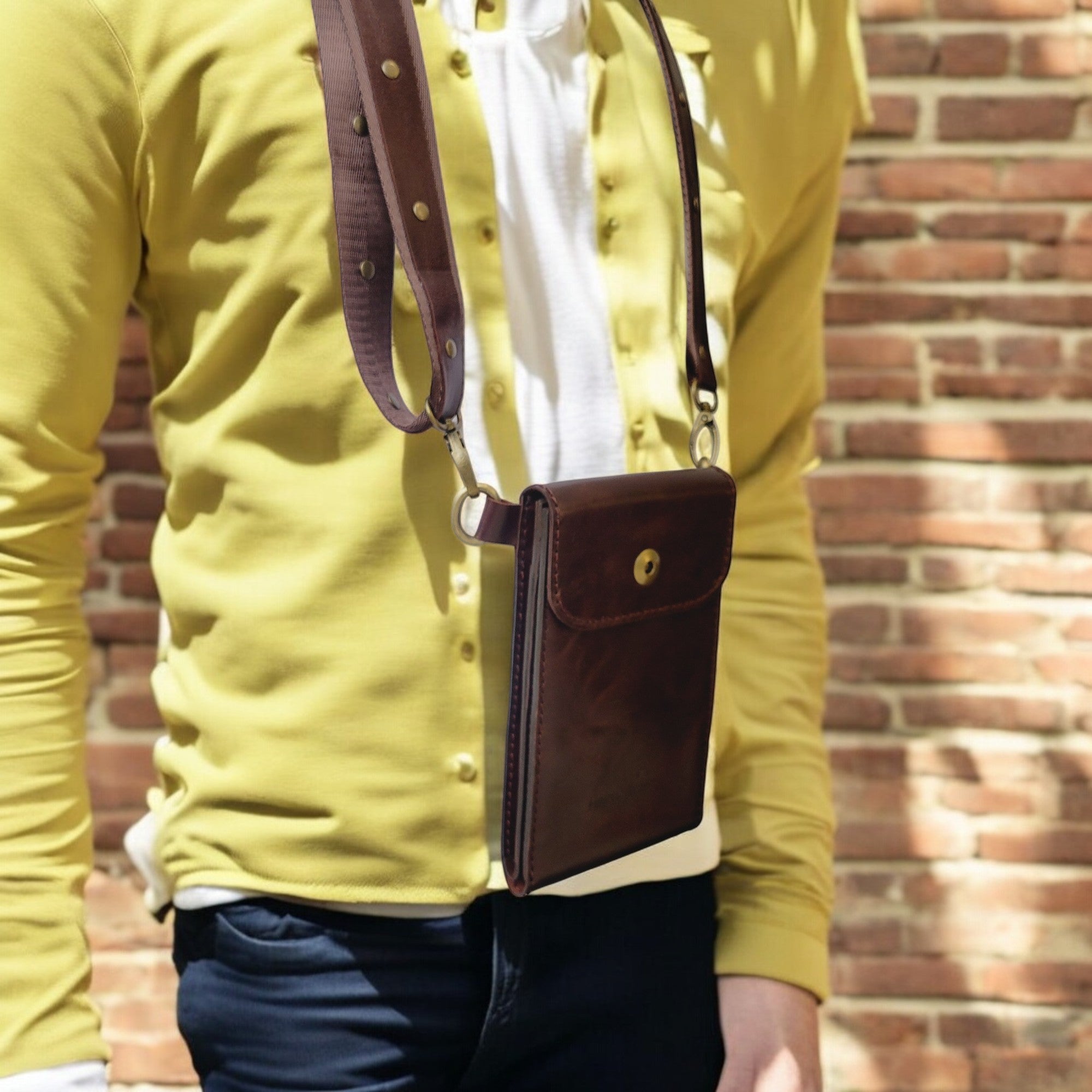 A person wearing a yellow shirt and dark pants is standing in front of a brick wall. The person is wearing a brown leather crossbody bag with an adjustable strap.