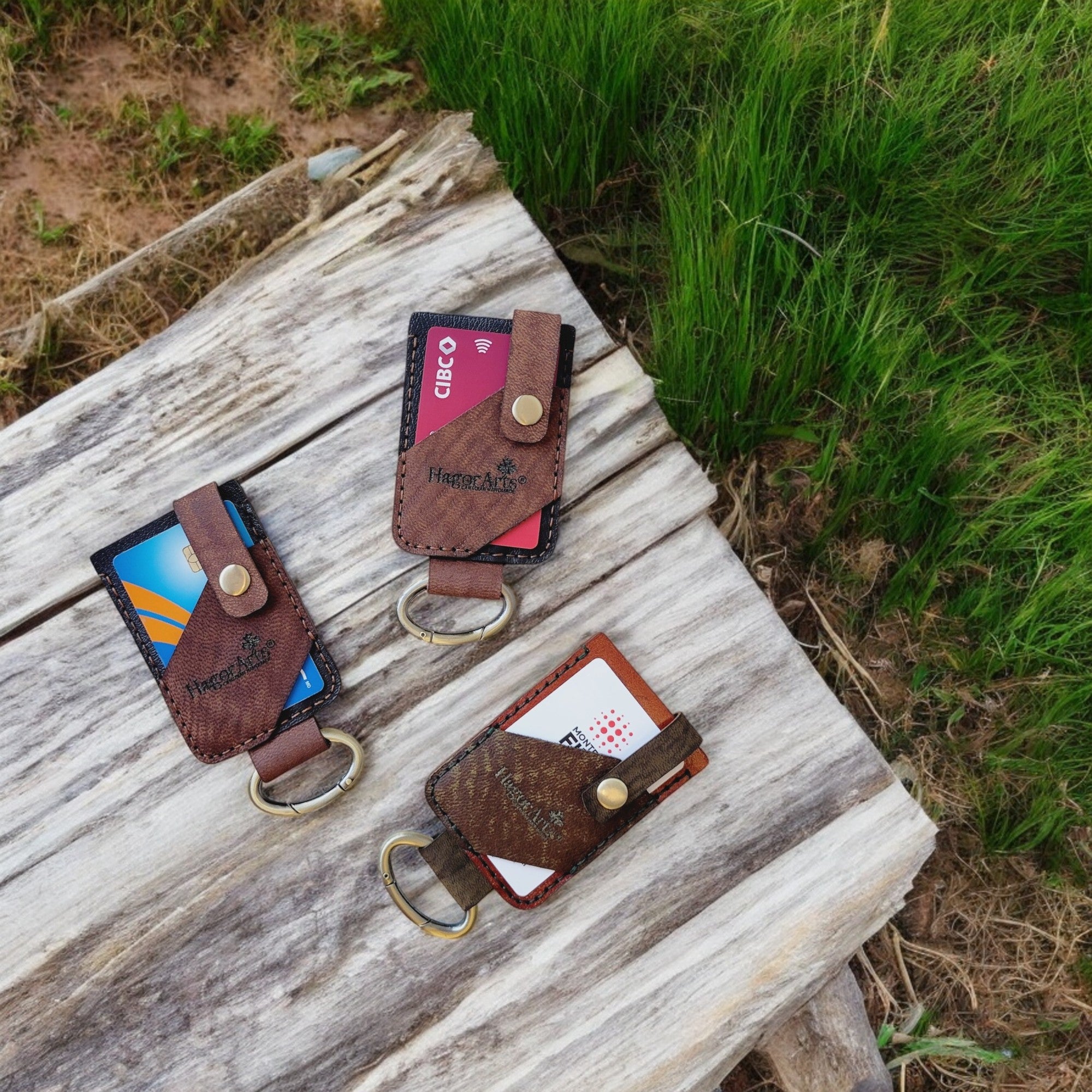 Three handmade leather card and key holders with a keychain attached, placed on a wooden surface with grass in the background.