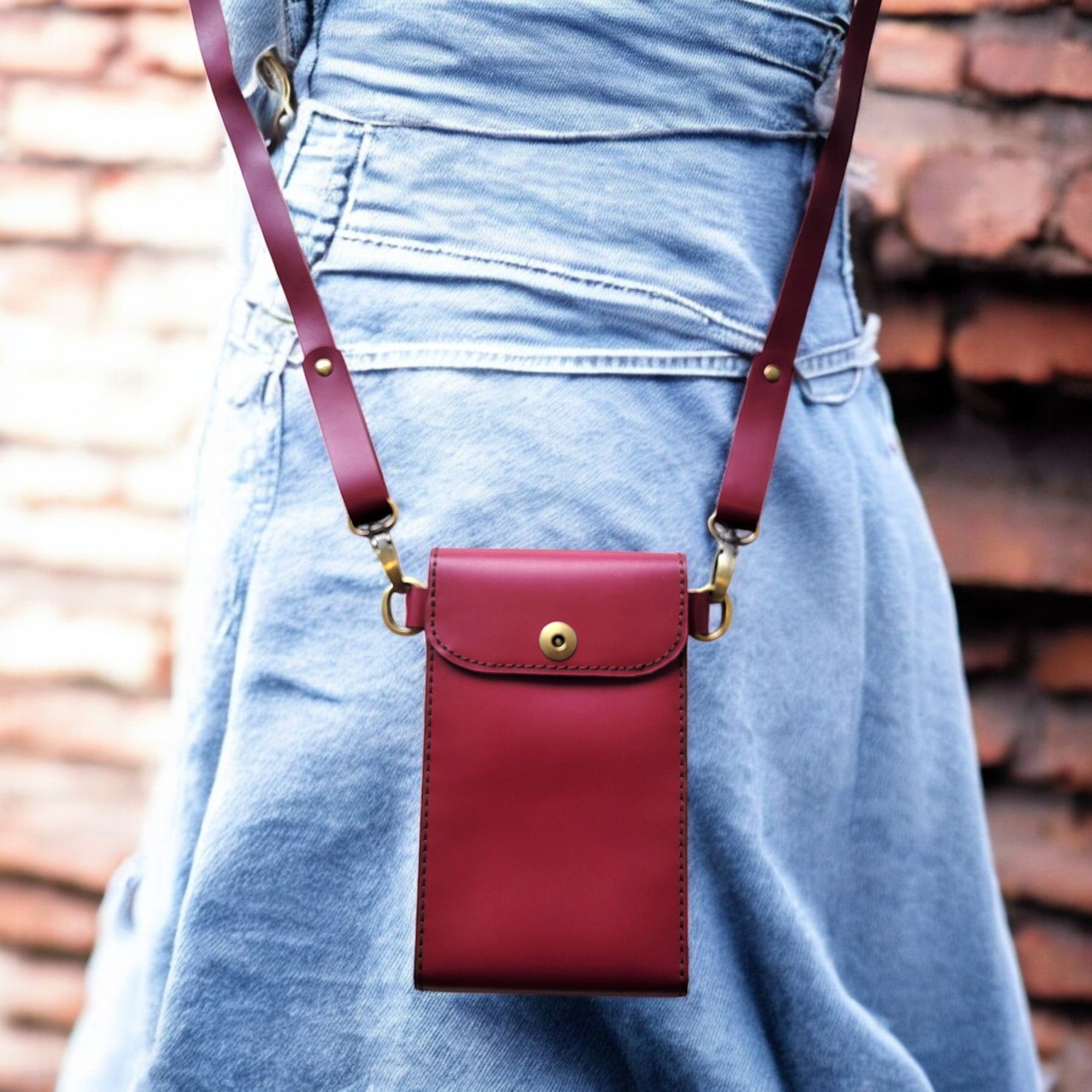A person wearing a blue denim skirt is carrying a claret red leather crossbody bag with an adjustable strap.