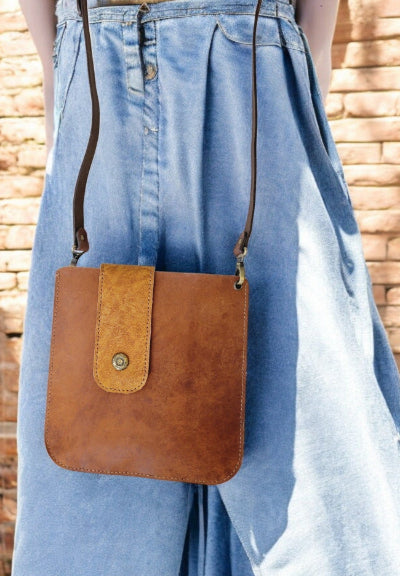 Brown leather crossbody bag worn by a person in blue jeans against a brick wall.