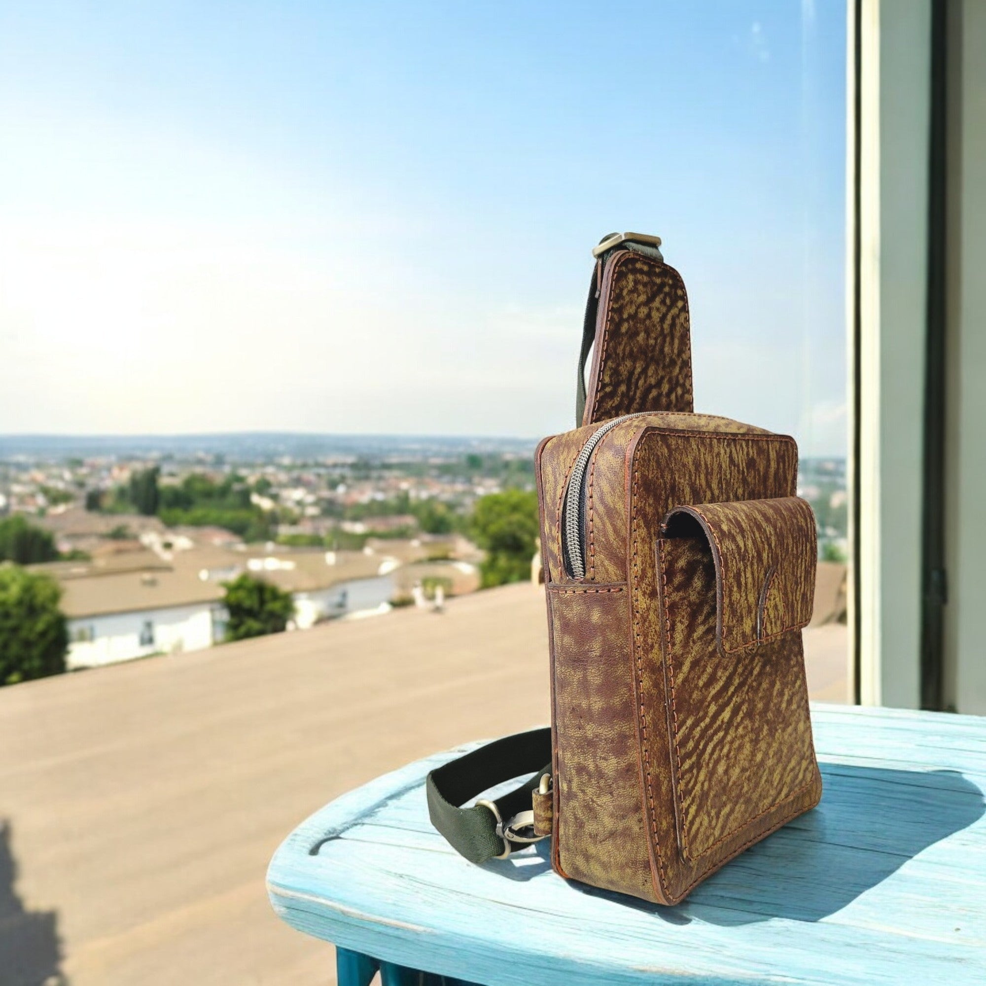 A brown full-grain leather sling bag with a textured finish, displayed on a table with a scenic background.
