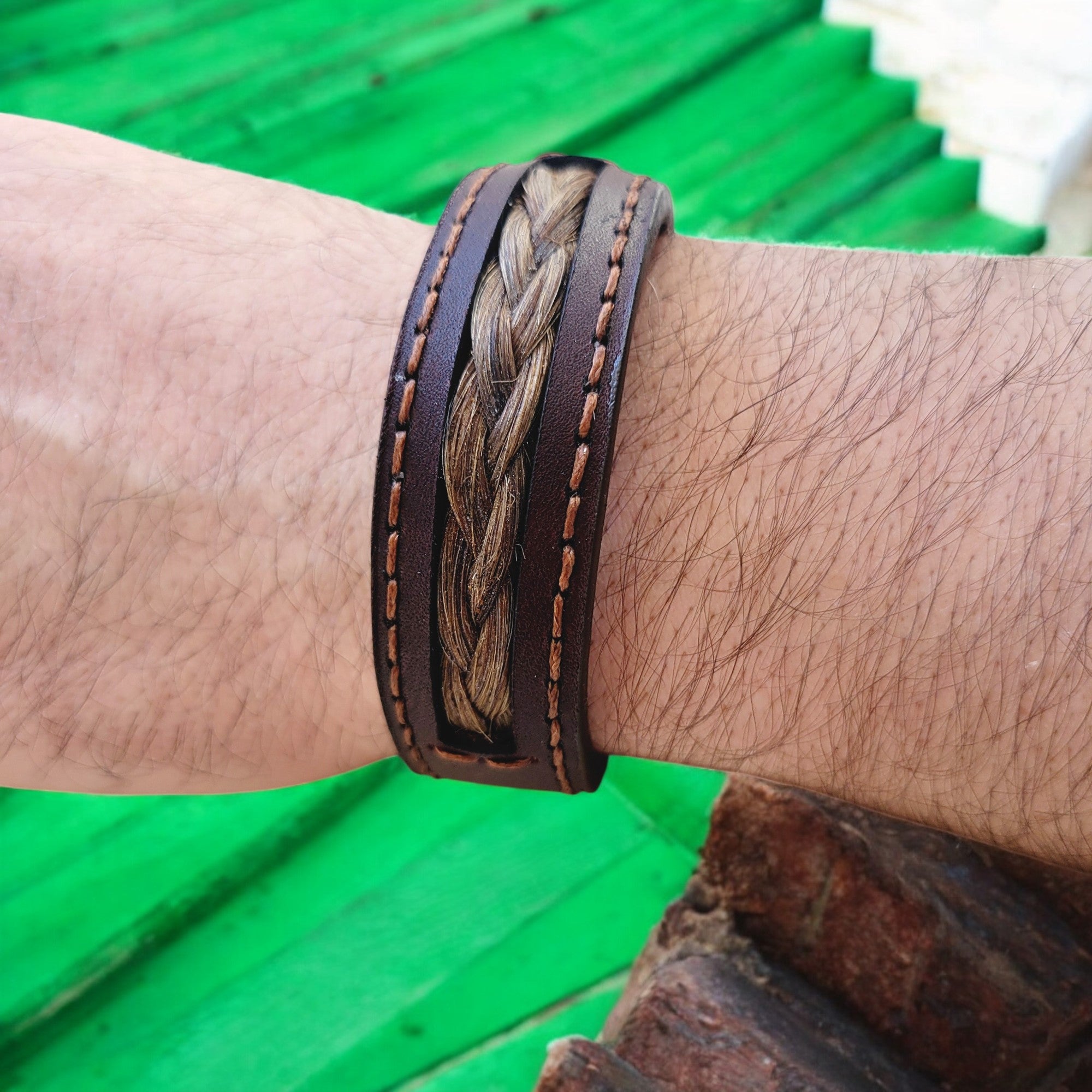 A person's wrist wearing a brown leather bracelet with a intricate design, featuring a strand of hair interwoven with the leather.