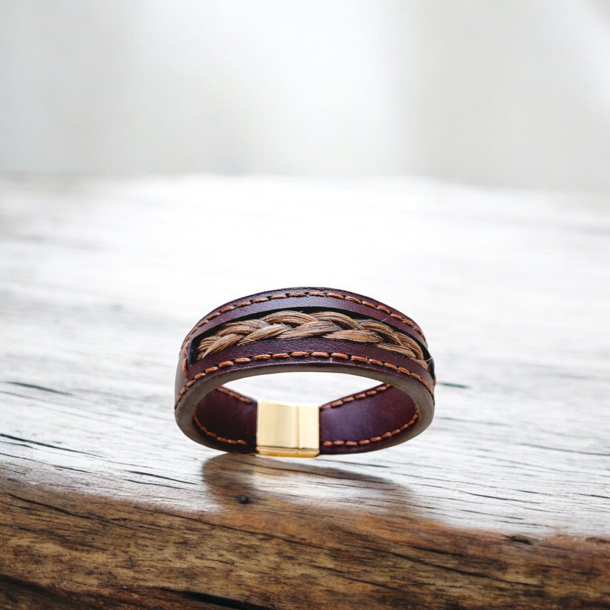 A brown leather bracelet with a woven pattern and a gold-tone clasp, displayed on a wooden surface.