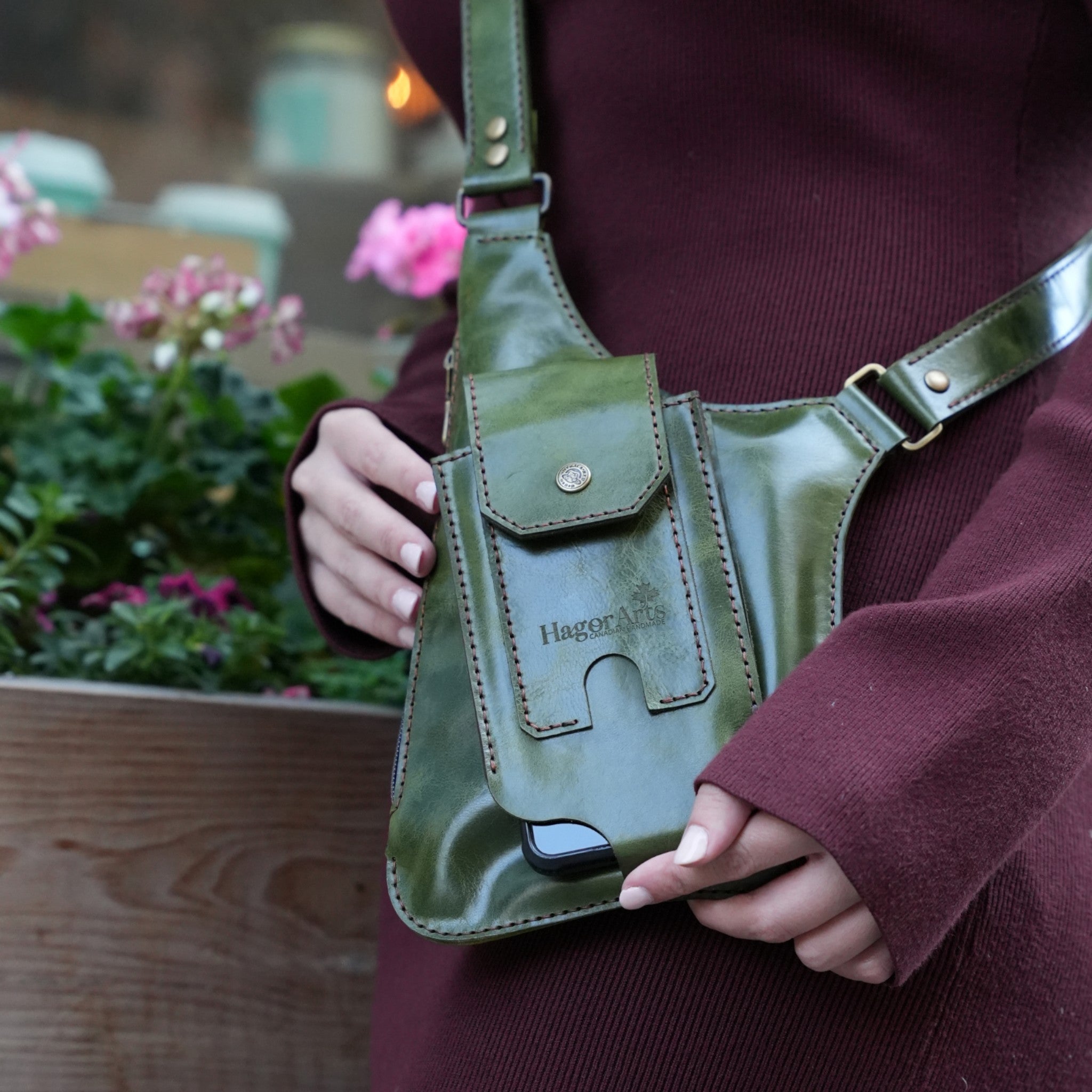 Person holding a green leather bag with 'Hagoromo' branding, outdoor setting with flowers.